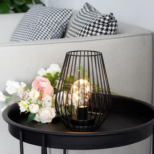 8'' cage-shaped battery lamp by JHY DESIGN with a black metal frame and glowing bulb on a round black table beside flowers.