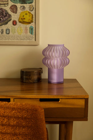 Oppi Table Lamp; purple ribbed lamp on a wooden desk beside a framed puzzle.