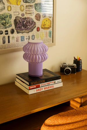 Oppi Table Lamp, purple ribbed base atop a stack of design books on a wooden desk