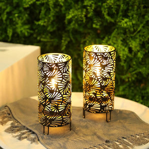Set of 2 9-inch metal lamps with leaf-cutout patterns; battery-powered cylindrical lanterns emitting warm glow.
