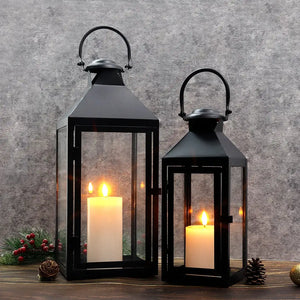 Set of 2 high black metal candle lanterns with glass panels and warm lit candles.