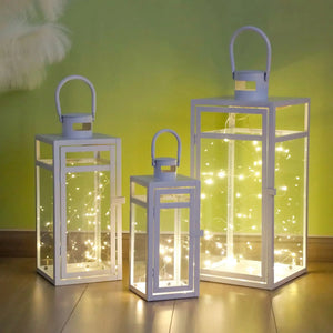 Set of 3 metal candle lanterns in blue-gray finish with glass panes and warm string lights.