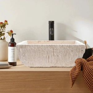 Rectangular light travertine bathroom sink with hand-chiseled exterior on a wooden vanity.