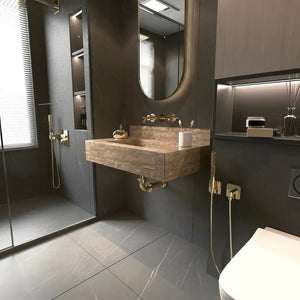 Bathroom travertine stone wall-mount sink, 24x16x5 in, beige stone with gold fixtures, round mirror, dark tiled walls.