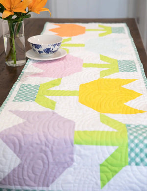 Tulip Quilted Table Runner Pattern by Block Party Quilt Co; vibrant fabric runner with tulip motifs and teal border.