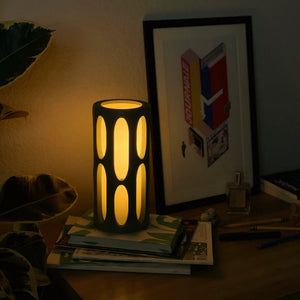 Amoeba table lamp by Terra Labs; black cylindrical base with oval cutouts, emitting warm ambient light on a desk.
