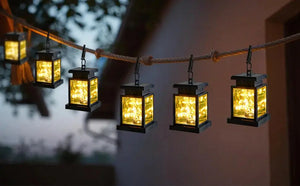 Set of 5 solar lantern lights with black metal frames and glass panels hung on a rope, emitting a warm yellow glow.