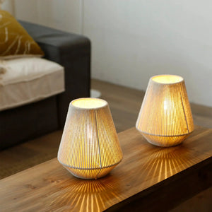 Set of 2 8-inch high woven, battery-operated table lamps with beige fabric shades and warm glow on a wooden table.