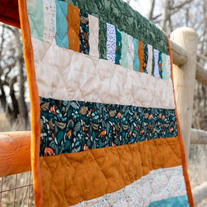 Bricks and Beams Quilt Kit - Timberline, patchwork quilt with teal, green and orange fabrics and orange binding.