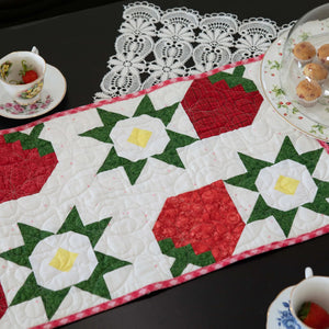 Strawberry table runner pattern; red and green patchwork with pink binding on white background