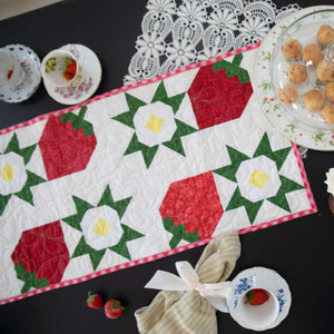Strawberry Table Runner Kit quilt on a black table with red-green patchwork and lace doily.
