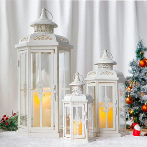 Set of three white metal candle lanterns with glass panels and gold brush detailing; heights 20, 14 and 10 inches, candles lit.