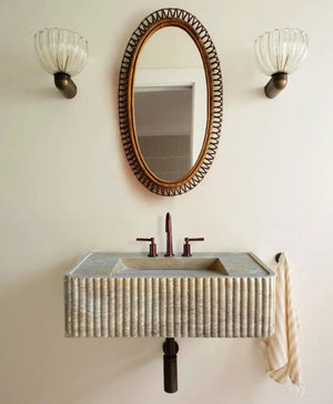 Walnut travertine rectangular wall-mount bathroom sink with fluted exterior; copper faucet, oval woven-frame mirror, and two sconces.