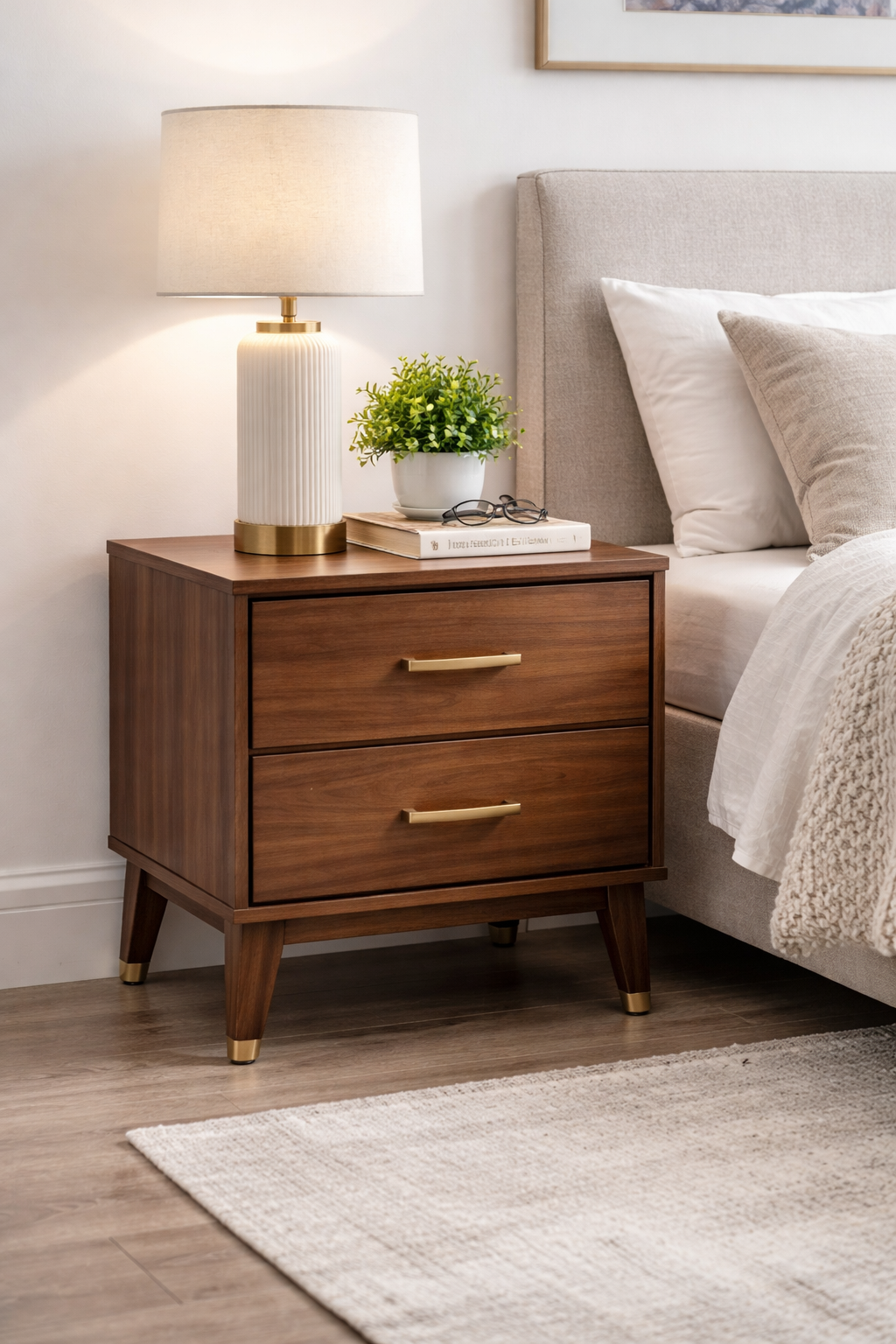 Modern wooden nightstand with two drawers and brass handles, styled beside a bed with a table lamp, books, and a small plant in a cozy bedroom setting.