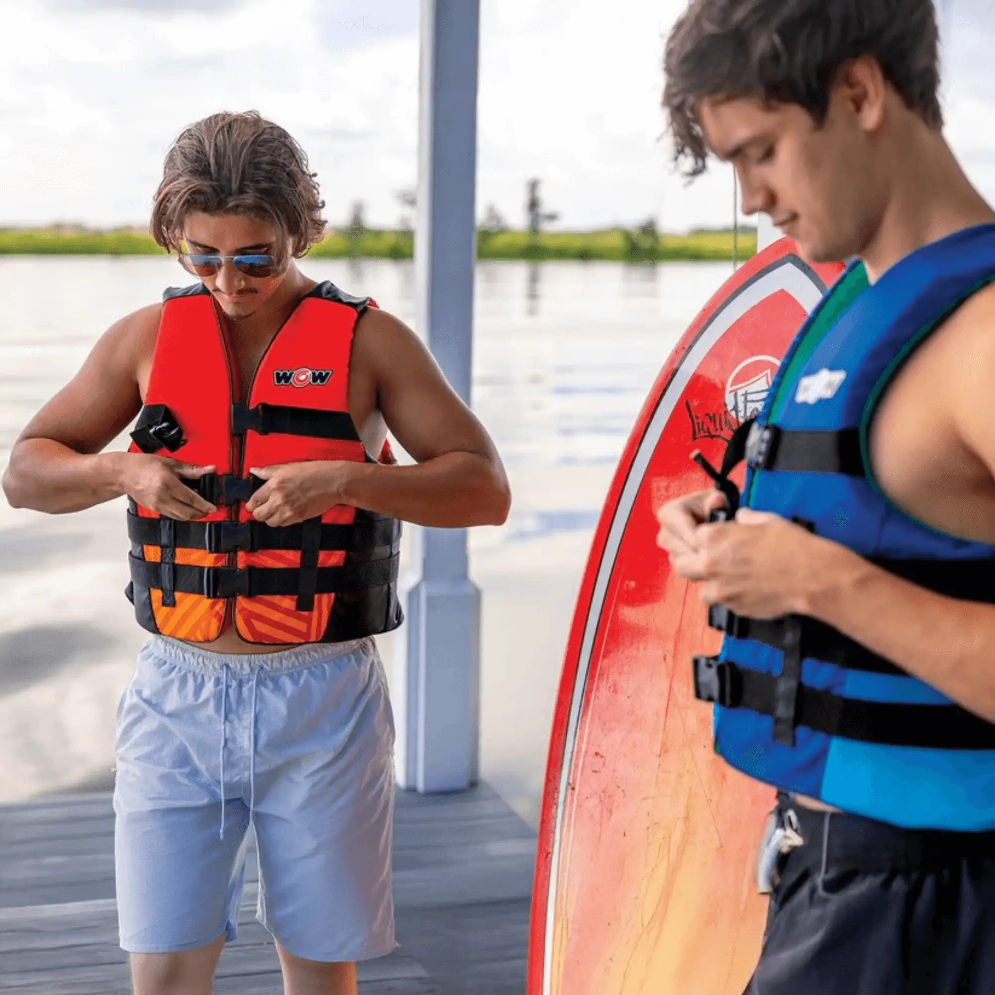 WOW Watersports Vis-Wave adult life vests; blue vest on the right and red/orange vest on the left by the water.