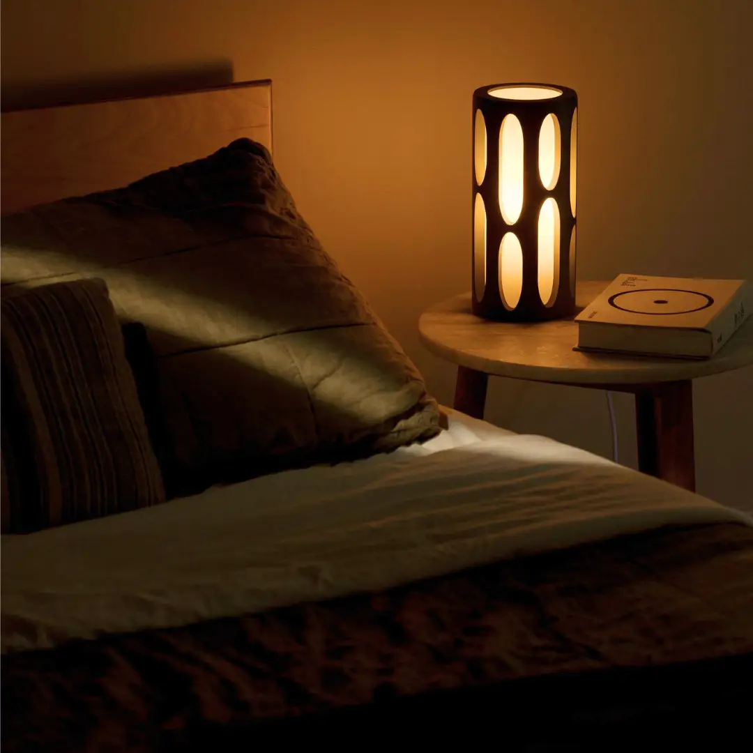 Amoeba Table Lamp by Terra Labs, black cylindrical lamp with oval cutouts, warm glow on bedside table.