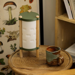 Toro Table Lamp by Terra Labs; green top, wooden posts, ribbed white glass body on a wooden table with two mugs nearby.