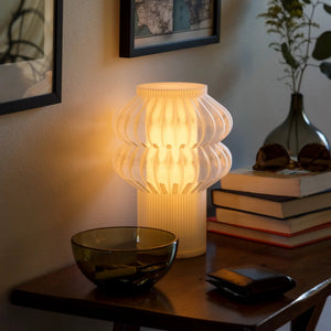 Oppi Table Lamp by Terra Labs; white ribbed ceramic shade glowing on a wooden table.