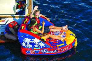 WOW Beach Bubba 2P towable raft in blue, red and yellow with palm tree graphics, two riders wearing life jackets.
