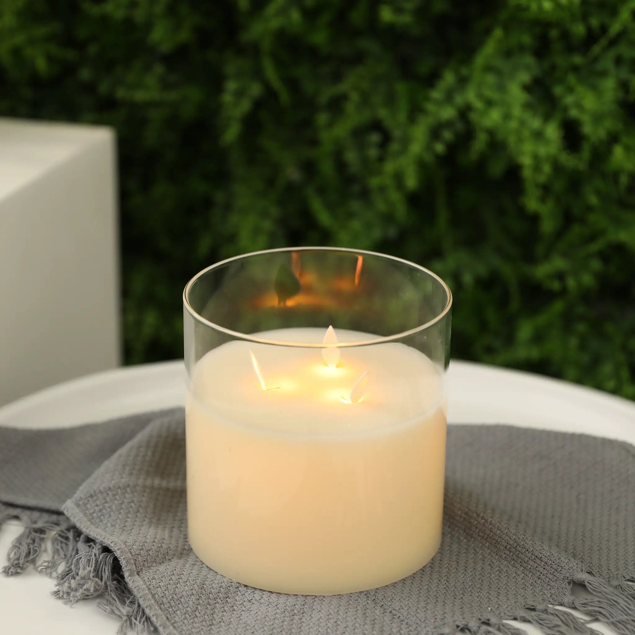 JHY DESIGN 6-inch glass 3-wick battery-operated flameless candle with warm glow on a gray woven cloth.