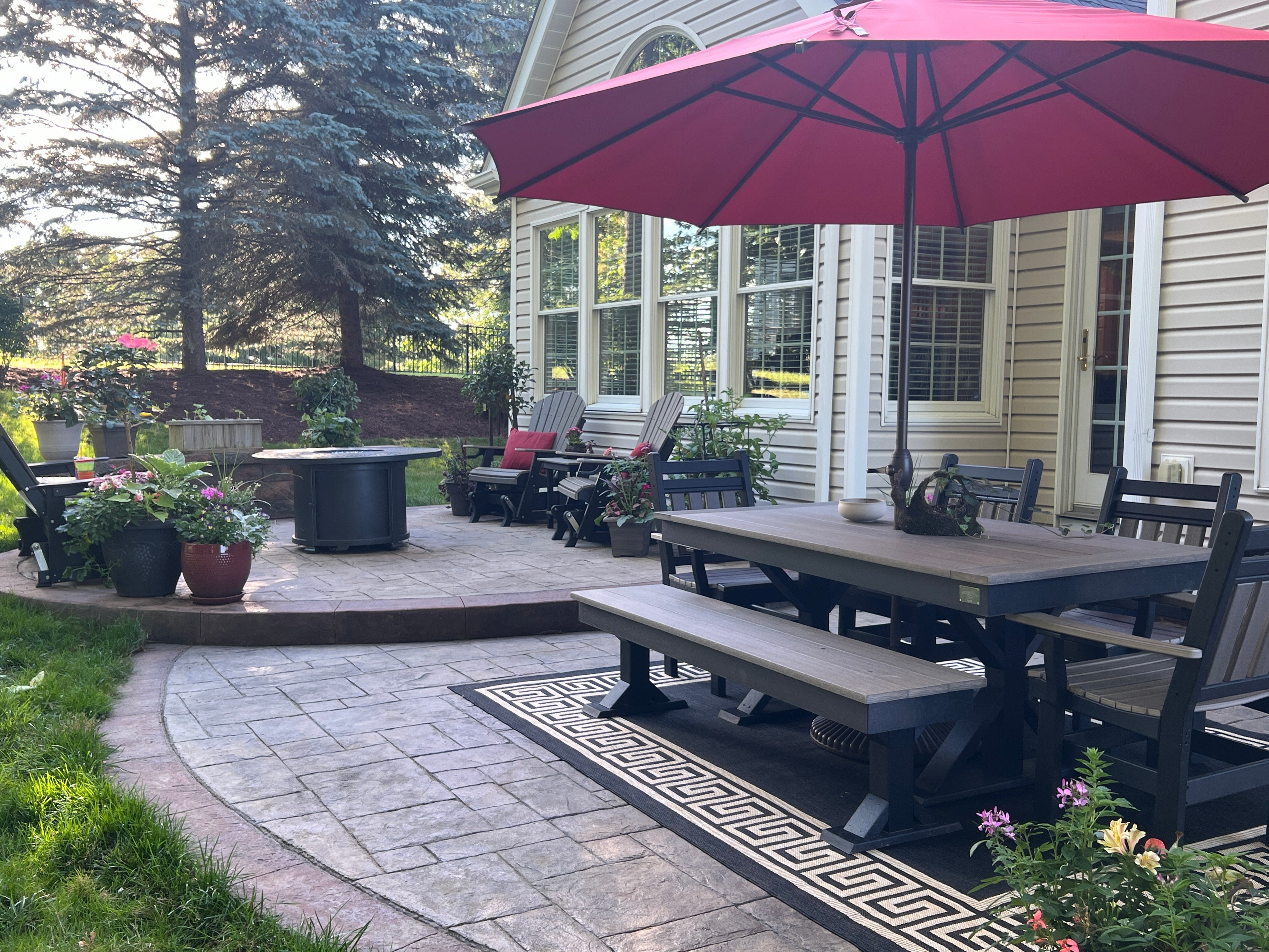 Trestle X Outdoor Bench in dark gray wood on a patio with a long dining table and pink umbrella