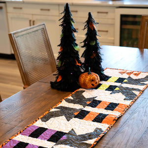 Witches Feet Table Runner Quilt Kit by Block Party Quilt Co; Halloween patchwork on a wooden table with two pumpkins.
