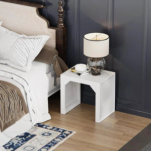 White slatted wood end table with glass lamp and books in a modern bedroom.