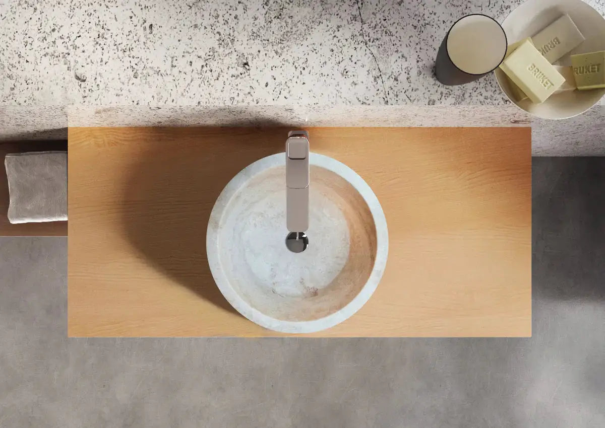 Troia Light travertine above-vanity sink; round light stone basin with chrome faucet on wood vanity.
