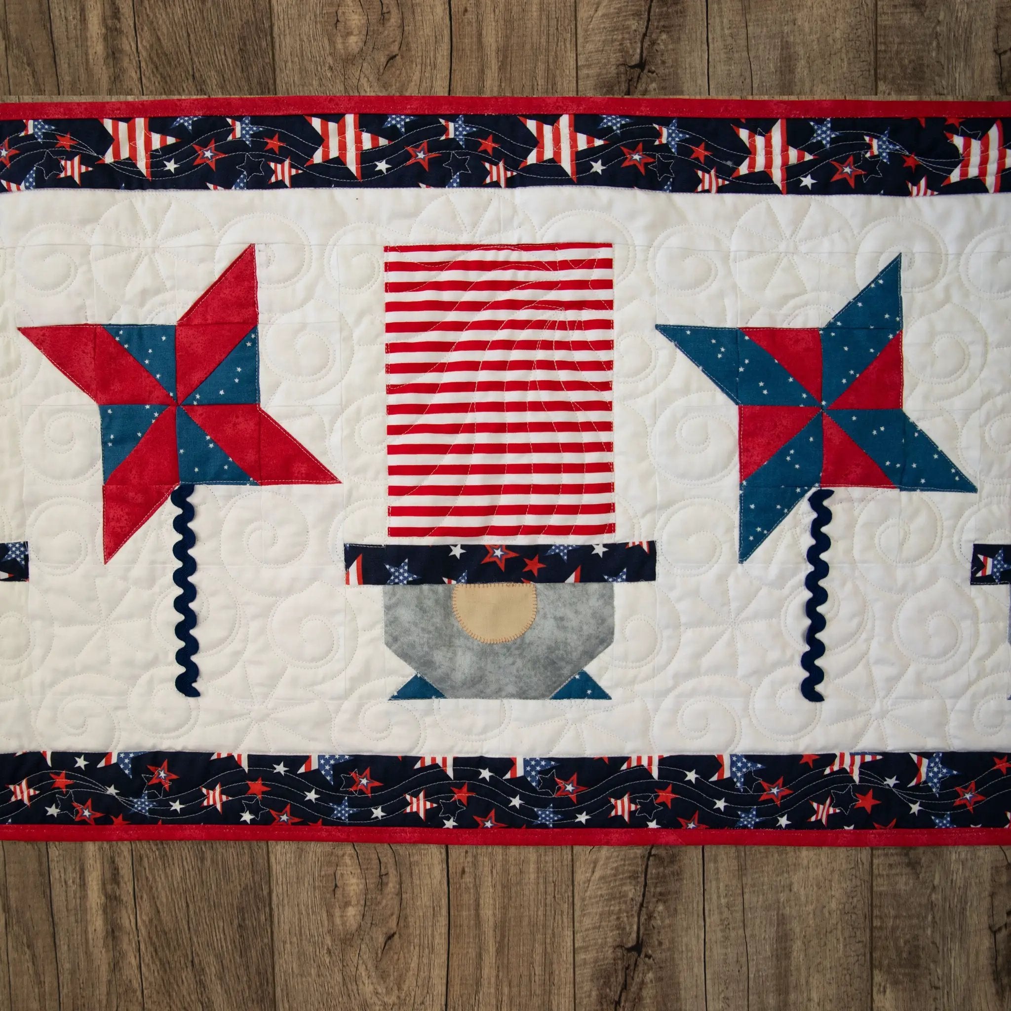 Patriotic Gnomes table runner quilt kit with red, white and blue star and stripe blocks on a white quilt background.