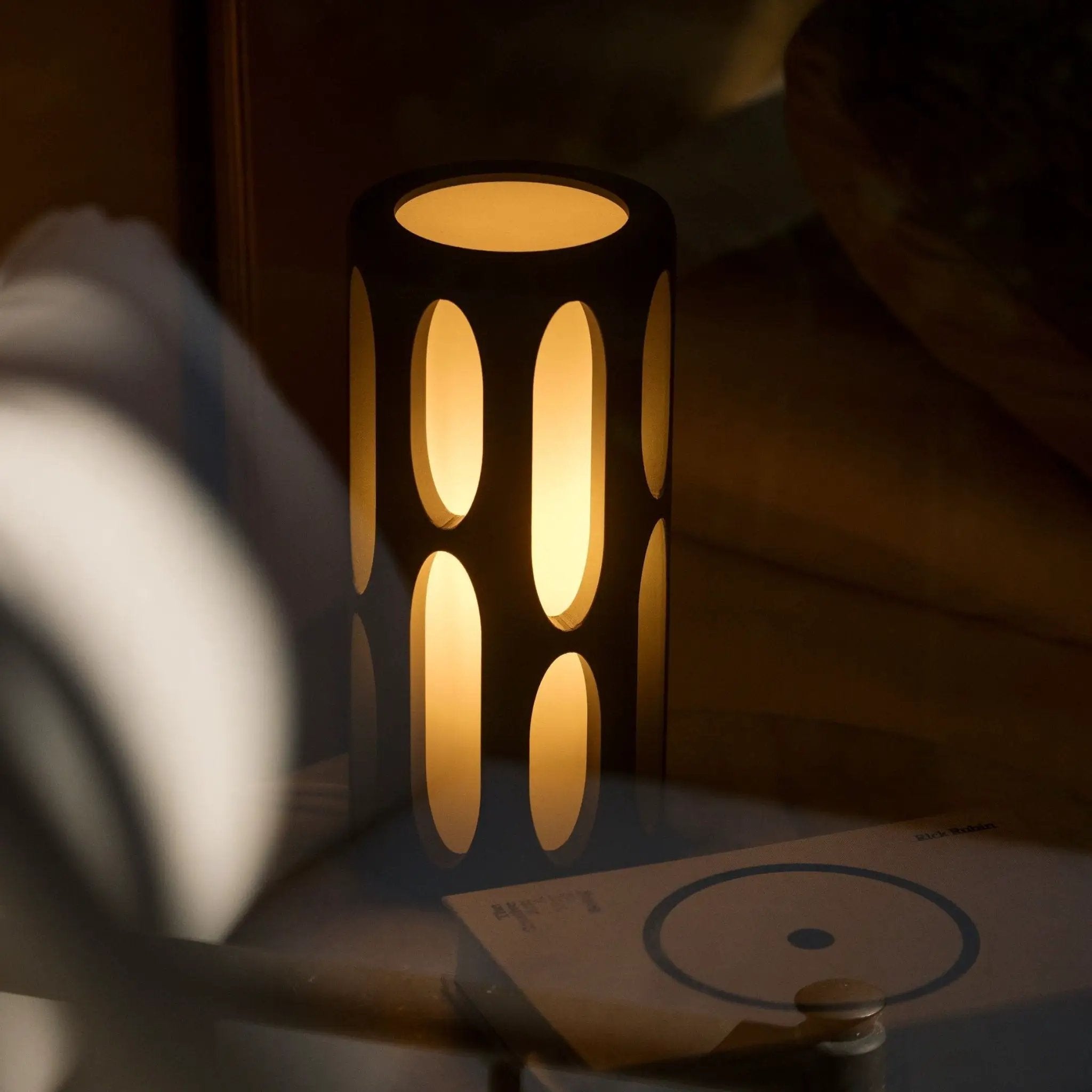 Amoeba Table Lamp by Terra Labs; black metal cylinder with large oval cutouts and warm amber glow.