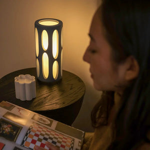 Amoeba Table Lamp by Terra Labs; dark cylindrical lamp with oval cutouts glowing warm amber on a wooden table.