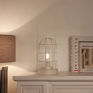 9.5-inch white metal cage table lamp, battery-operated, on a mantel beside books