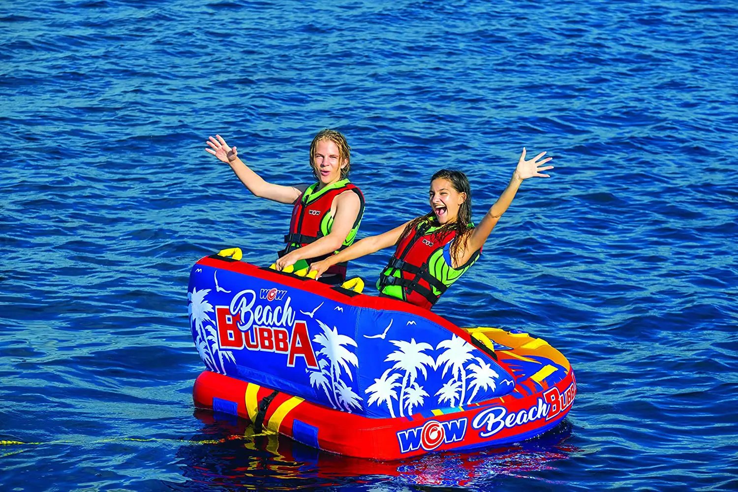 WOW Beach Bubba 2-person towable raft; blue with palm tree graphics, red trim, two riders in life jackets on blue water.