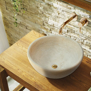 Beige travertine above-counter vessel sink on wood vanity with rose-gold faucet, stone wall backdrop.