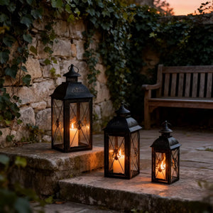 Set of four black metal decorative lanterns with glass panels and candlelight along a stone patio.