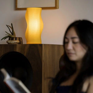 Woogie table lamp by Terra Labs, yellow ribbed curved shade on a wooden shelf.