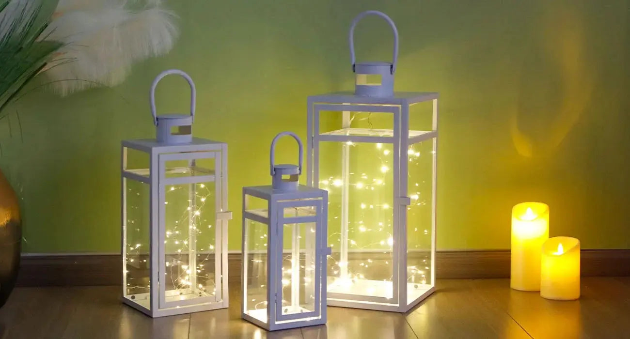 Set of 3 white metal candle lanterns with glass panels and handles in varying heights, lit by string lights.