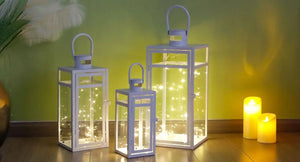 Set of 3 white metal candle lanterns with glass panels and handles in varying heights, lit by string lights.