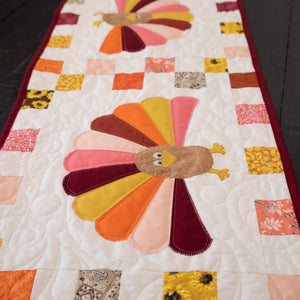 Quilt kit turkey table runner featuring multicolor tail feathers, patchwork squares, and burgundy binding