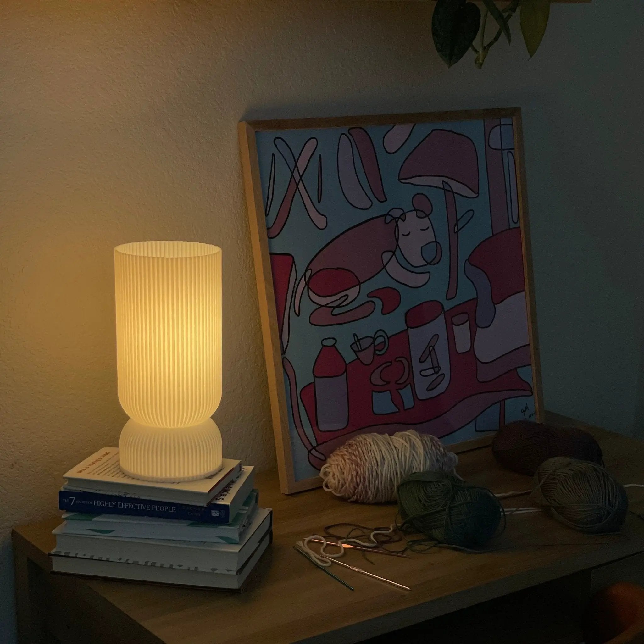 Stance table lamp with ribbed ivory shade and base, resting on a stack of books beside a colorful framed artwork.