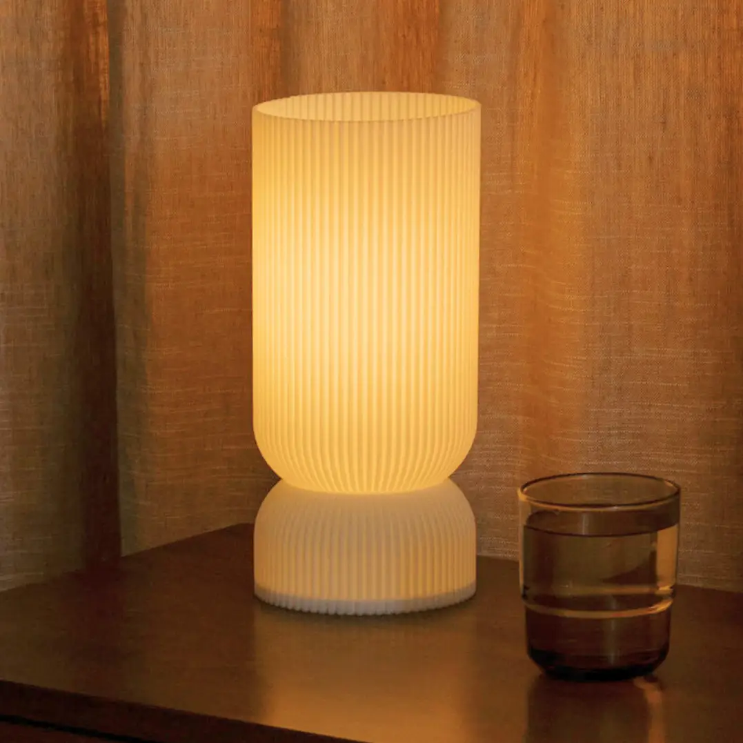 Stance table lamp, white ribbed shade with warm glow on a dark wooden surface beside 1 glass tumbler.