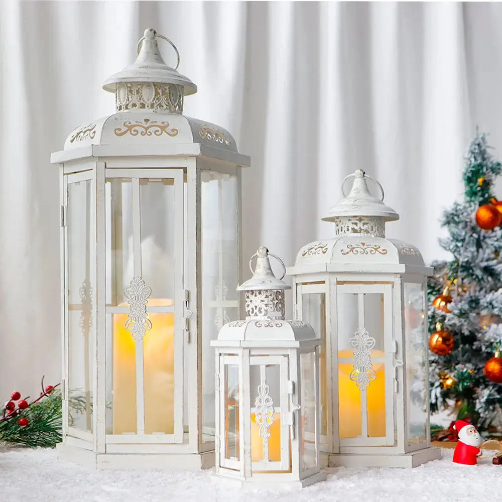 Set of three white metal candle lanterns with glass panels and gold brush detailing; heights 20, 14 and 10 inches, candles lit.
