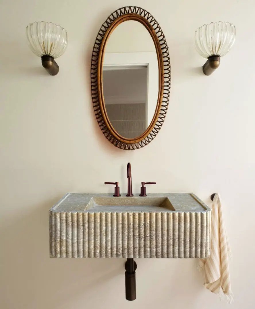 Walnut travertine rectangular wall-mount bathroom sink with fluted exterior; copper faucet, oval woven-frame mirror, and two sconces.