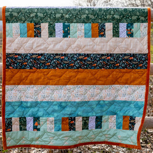 Bricks and Beams Quilt Kit Timberline by Block Party Quilt Co; vibrant patchwork with teal greens and orange binding.