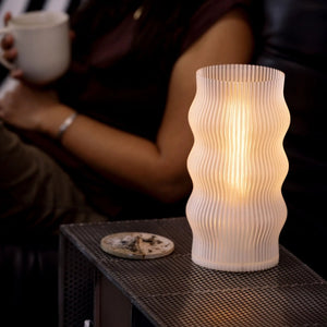 Yuji table lamp by Terra Labs; white ribbed glass base with warm glow on a metal mesh shelf.