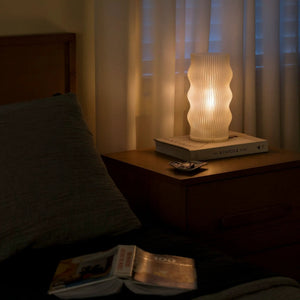 Yuji table lamp by Terra Labs with a ribbed white body on a bedside table, emitting warm light.